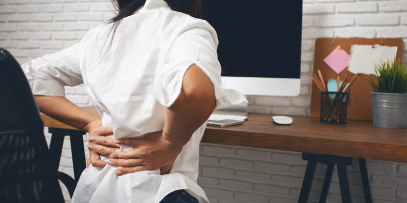 A woman holding her back in pain while working from home on her laptop.