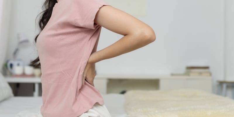 A woman experiencing lower back pain while sitting on a bed, used to explain if bed rest is good for back pain or if it hinders recovery.
